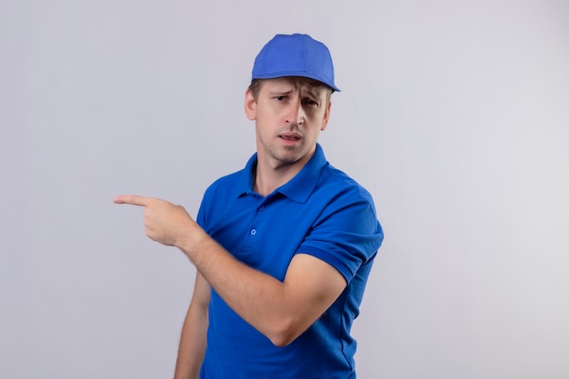 Repartidor guapo joven en uniforme azul y gorra apuntando con el dedo índice hacia el lado mirando confundido parado sobre pared blanca