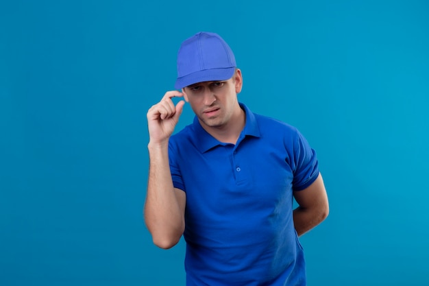 Repartidor guapo joven en uniforme azul y gorra disgustado tocando su gorra con el ceño fruncido de pie sobre la pared azul