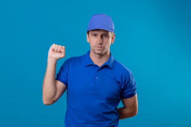 Repartidor guapo joven en uniforme azul y gorra levantando el puño con expresión seria y segura en la cara de pie sobre la pared azul