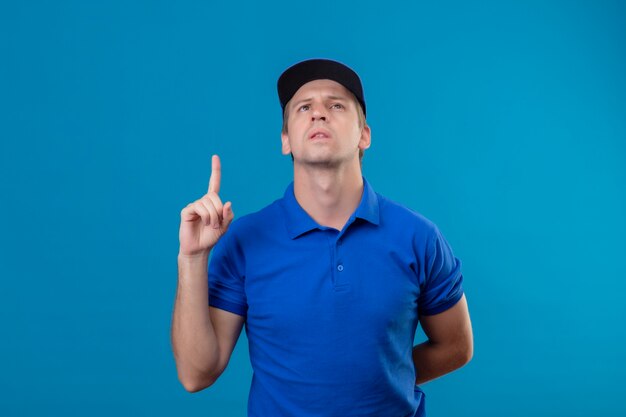 Repartidor guapo joven en uniforme azul y gorra mirando hacia arriba apuntando con el dedo hacia arriba recordándose a sí mismo que no debe olvidar lo importante que está parado sobre la pared azul