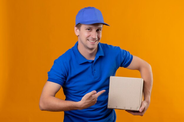 Repartidor guapo joven en uniforme azul y gorra con paquete de caja apuntando con el dedo a él