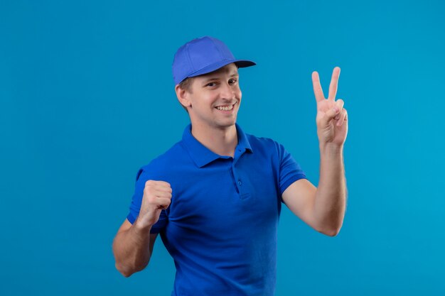 Repartidor guapo joven en uniforme azul y gorra que muestra el número dos sonriendo alegremente de pie sobre la pared azul