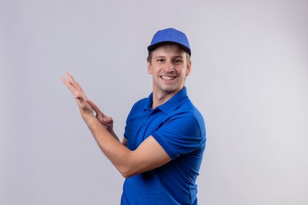 Repartidor guapo joven en uniforme azul y gorra sonriendo amistosamente apuntando con el dedo al brazo de su mano de pie sobre la pared blanca