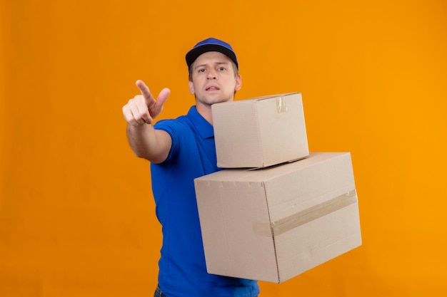 Repartidor guapo joven en uniforme azul y gorra sosteniendo cajas de cartón apuntando a algo con el dedo