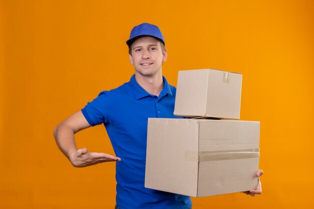 Repartidor guapo joven en uniforme azul y gorra sosteniendo cajas de cartón que presentan con el brazo de su mano de pie sobre la pared naranja