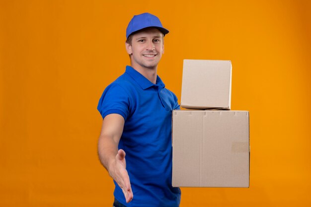 Repartidor guapo joven en uniforme azul y gorra sosteniendo cajas de cartón sonriendo saludo amistoso ofreciendo mano de pie sobre la pared naranja