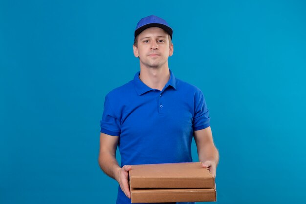 Repartidor guapo joven en uniforme azul y gorra sosteniendo cajas de pizza con expresión de confianza en la cara de pie sobre la pared azul