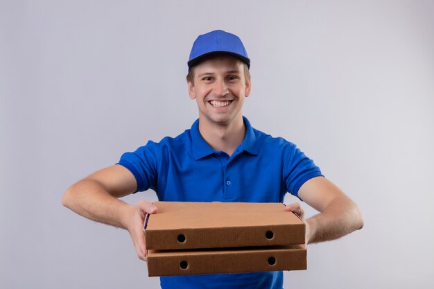 Repartidor guapo joven en uniforme azul y gorra sosteniendo cajas de pizza sonriendo amigable de pie sobre la pared blanca