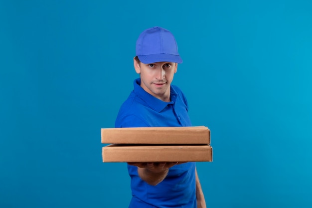 Repartidor guapo joven en uniforme azul y gorra sosteniendo cajas de pizza con sonrisa de confianza en la cara de pie sobre la pared azul