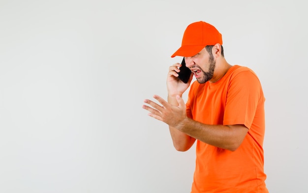 Repartidor hablando por teléfono móvil en camiseta naranja, gorra y con enojo. vista frontal.