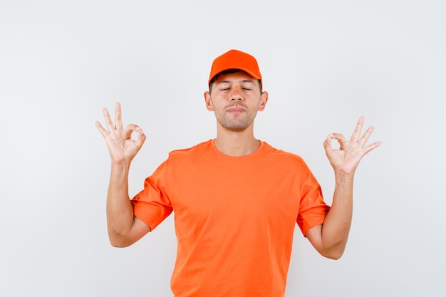 Foto gratuita repartidor haciendo bien firmar con los ojos cerrados en camiseta naranja y gorra y mirando pacífico