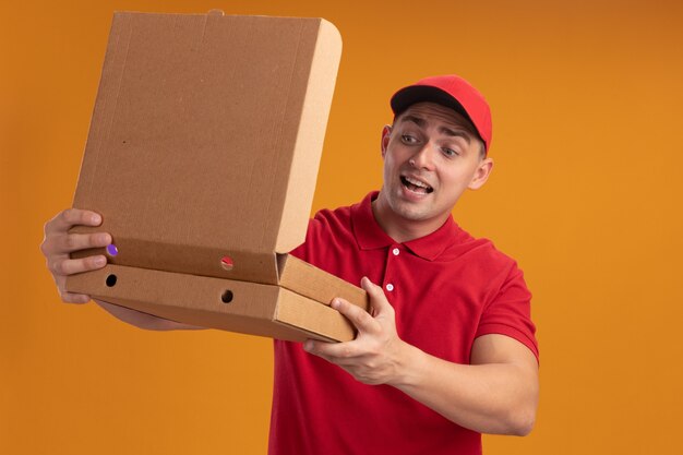 Repartidor joven alegre vestido con uniforme con apertura de tapa y mirando la caja de pizza aislada en la pared naranja