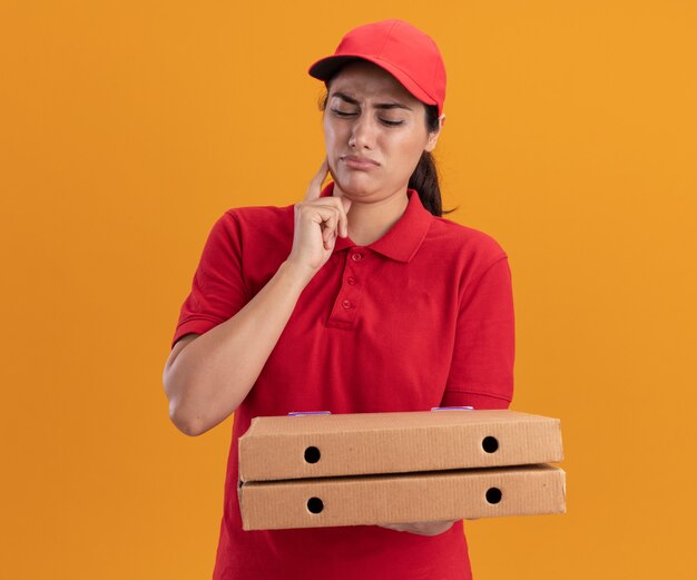 Repartidor joven aprensivo con uniforme y gorra sosteniendo y mirando cajas de pizza poniendo el dedo en la mejilla aislado en la pared naranja