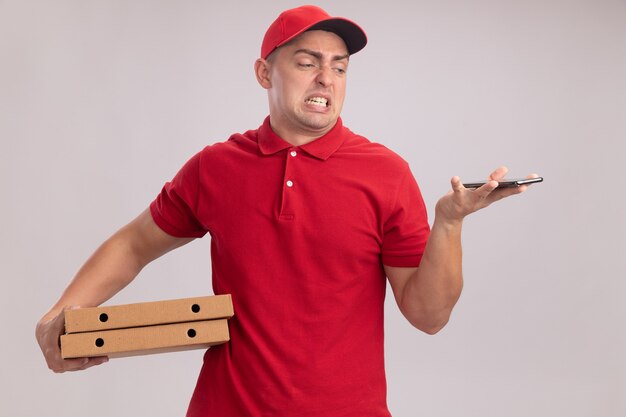 Repartidor joven aprensivo vestido con uniforme con gorra sosteniendo cajas de pizza y mirando el teléfono en la mano aislado en la pared blanca