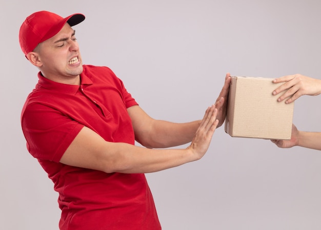 Repartidor joven asustado vestido con uniforme con gorra dando caja al cliente aislado en la pared blanca