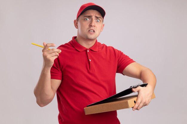 Repartidor joven asustado vestido con uniforme con gorra sosteniendo caja de pizza con portapapeles aislado en la pared blanca