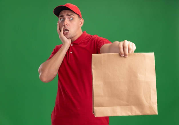 Repartidor joven asustado vestido con uniforme y gorra sosteniendo el paquete de comida de papel en la cámara poniendo la mano en la mejilla aislada en la pared verde