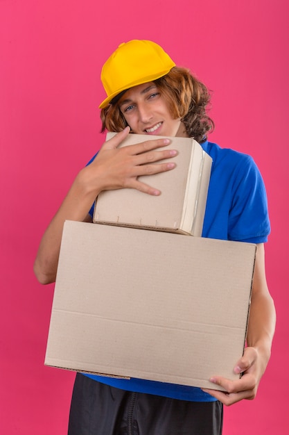 Repartidor joven con camisa polo azul y gorra amarilla sosteniendo cajas de cartón soñando abrazando cajas sonriendo con cara feliz sobre fondo rosa aislado