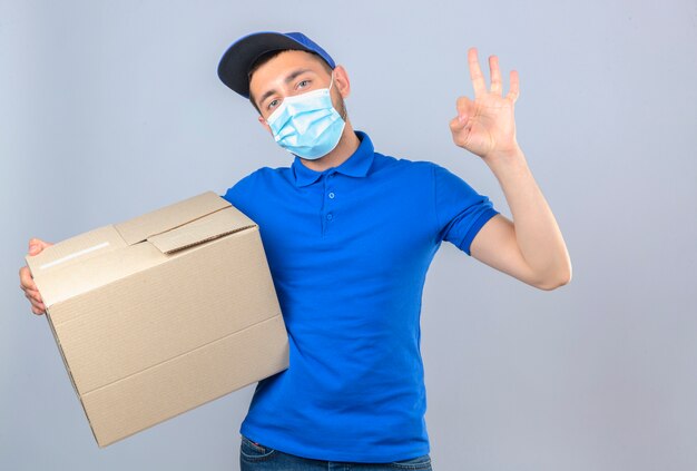 Repartidor joven con camisa polo azul y gorra en máscara protectora médica de pie con caja de cartón y haciendo bien firmar sobre fondo blanco aislado