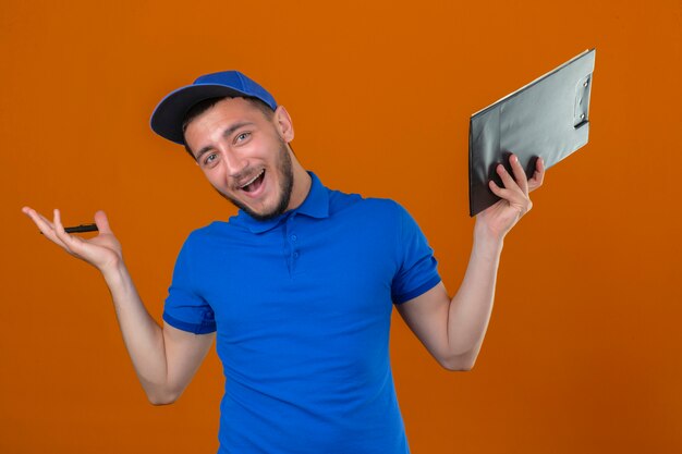 Repartidor joven con camisa polo azul y gorra de pie con portapapeles con sonrisa feliz y expresión de ganador con la mano levantada sobre fondo naranja aislado