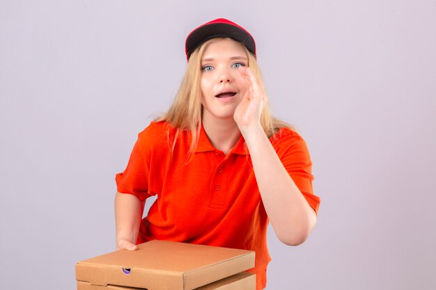 Repartidor joven en camisa polo naranja y gorra roja de pie con cajas de pizza sosteniendo la mano cerca de la boca abierta y diciendo algo sobre fondo blanco aislado