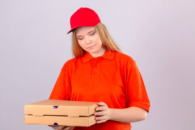 Repartidor joven en camisa polo naranja y gorra roja sosteniendo cajas de pizza mirando hacia abajo con una sonrisa en la cara sobre fondo blanco aislado