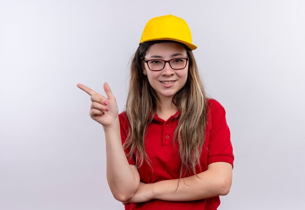 Repartidor joven en camisa polo roja y amarillo amistoso sonriente apuntando con el dedo hacia el lado