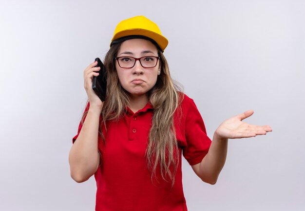 Repartidor joven en camisa polo roja y gorra amarilla hablando por teléfono móvil mirando confundido encogiéndose de hombros