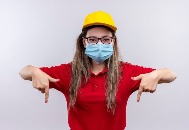 Foto gratuita repartidor joven en camisa polo roja y gorra amarilla con máscara facial protectora apuntando con el dedo índice hacia abajo