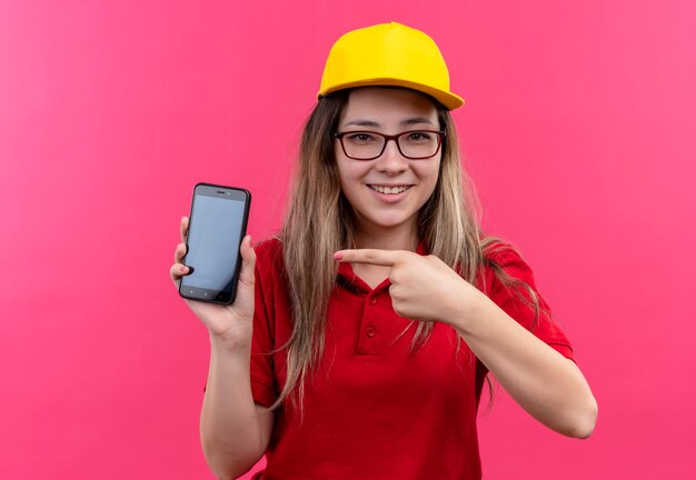 Repartidor joven en camisa polo roja y gorra amarilla mostrando smartphone apuntando con el dedo índice sonriendo