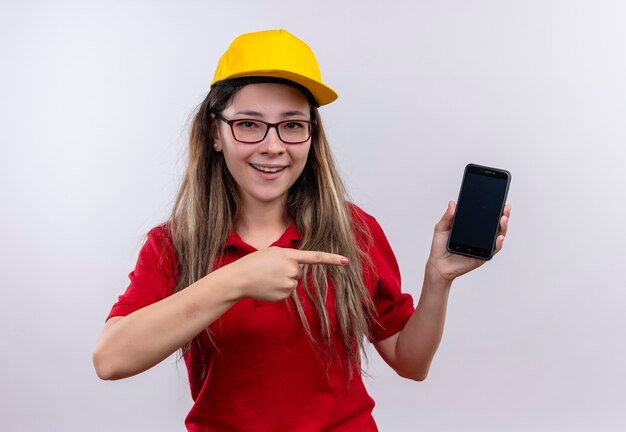 Repartidor joven en camisa polo roja y gorra amarilla mostrando smartphone apuntando con el dedo sonriendo alegremente