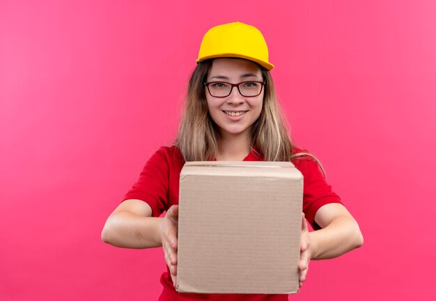 Foto gratuita repartidor joven en camisa polo roja y gorra amarilla con paquete de caja mirando a la cámara sonriendo confiado