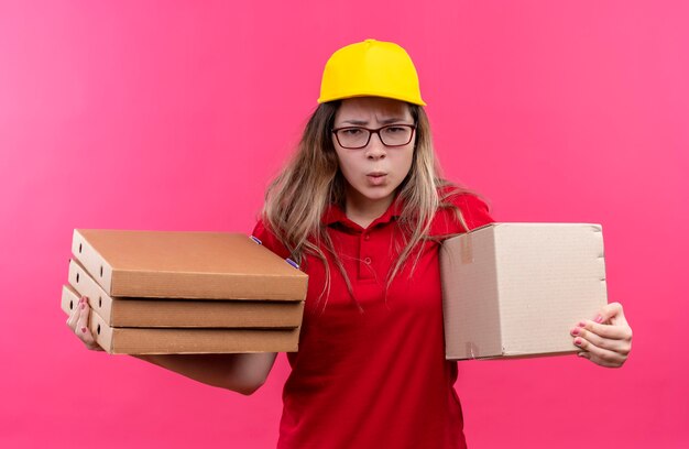 Repartidor joven en camisa polo roja y gorra amarilla con pila de cajas de pizza y paquete de caja mirando cametra con el ceño fruncido