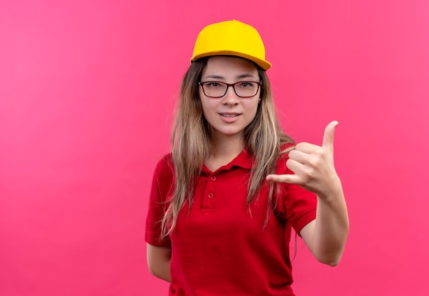 Repartidor joven en camisa polo roja y gorra amarilla sonriendo haciendo gesto de llamarme