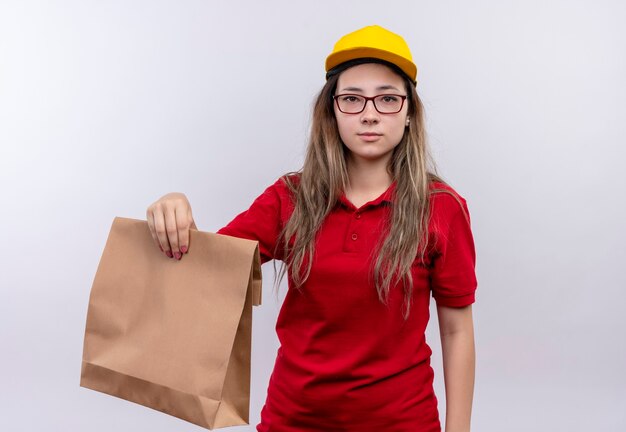 Repartidor joven en camisa polo roja y gorra amarilla sosteniendo el paquete de papel mirando a la cámara con expresión seria y segura