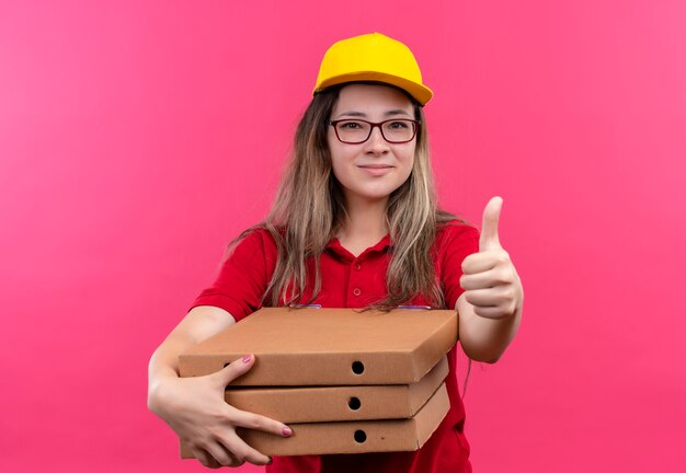 Repartidor joven en camisa polo roja y gorra amarilla sosteniendo una pila de cajas de pizza mirando a la cámara con una sonrisa de confianza en la cara mostrando los pulgares para arriba