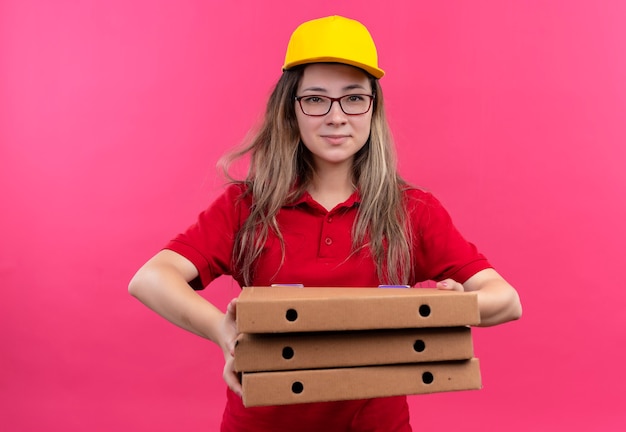 Repartidor joven en camisa polo roja y gorra amarilla sosteniendo una pila de cajas de pizza mirando a la cámara con una sonrisa de confianza en la cara