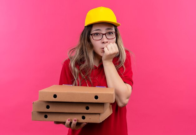 Repartidor joven en camisa polo roja y gorra amarilla sosteniendo una pila de cajas de pizza preocupado mordiendo las uñas