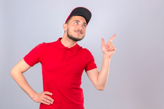 Repartidor joven con camisa polo roja y gorra apuntando con el dedo hacia arriba en el espacio de la copia mirando con una sonrisa en la cara sobre fondo blanco aislado