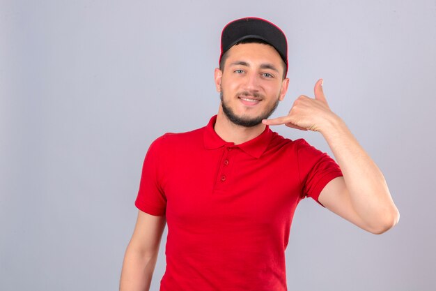 Repartidor joven con camisa polo roja y gorra haciendo gesto de llamarme mirando confiado sobre fondo blanco aislado