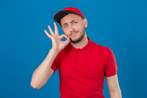 Repartidor joven con camisa polo roja y gorra haciendo gesto de silencio haciendo como cerrar la boca con una cremallera sobre fondo azul aislado