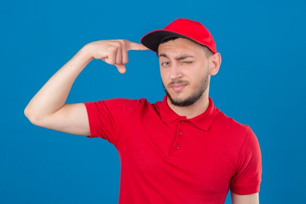 Repartidor joven con camisa polo roja y gorra mirando a la cámara guiñando un ojo apuntando con el dedo índice a la cabeza mirando inteligente sobre fondo azul aislado