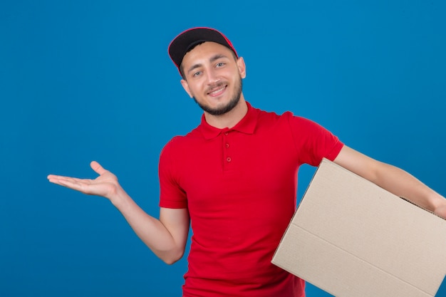 Repartidor joven con camisa polo roja y gorra de pie con caja sonriendo alegre presentando y apuntando con la palma de la mano mirando a la cámara sobre fondo azul aislado