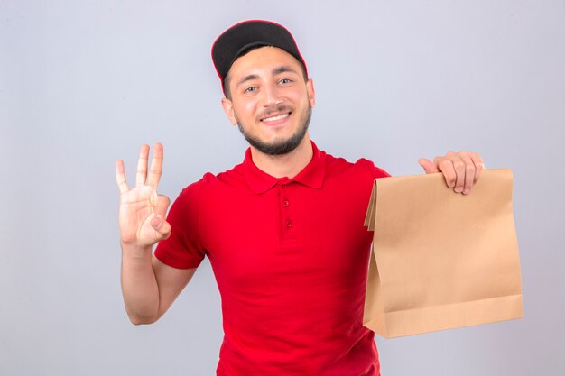 Repartidor joven con camisa polo roja y gorra de pie con el paquete de papel mostrando y apuntando hacia arriba con los dedos número tres mientras sonríe confiado y feliz sobre fondo blanco aislado