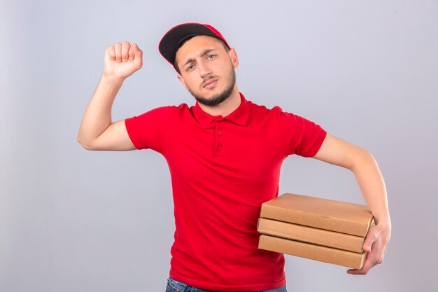 Repartidor joven con camisa polo roja y gorra de pie con una pila de cajas de pizza mostrando el puño a la cámara expresión facial agresiva sobre fondo blanco aislado