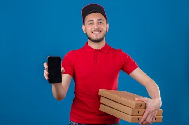 Foto gratuita repartidor joven con camisa polo roja y gorra de pie con pila de cajas de pizza y mostrando smartphone sonriendo amigable mirando a cámara sobre fondo azul aislado