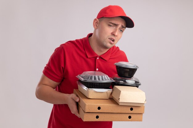 Repartidor joven cansado vestido con uniforme con tapa sosteniendo contenedores de comida en cajas de pizza aisladas en la pared blanca