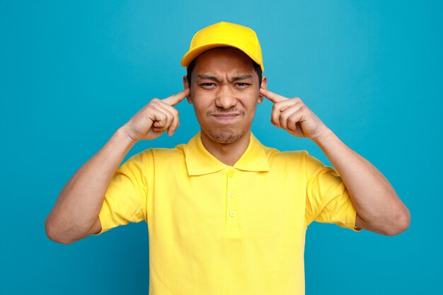 Repartidor joven con el ceño fruncido con uniforme y gorra manteniendo los dedos en las orejas