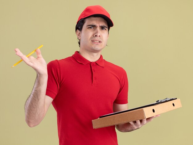 Repartidor joven con el ceño fruncido en uniforme rojo y gorra con portapapeles de paquete de pizza y lápiz mirando al frente aislado en la pared verde oliva