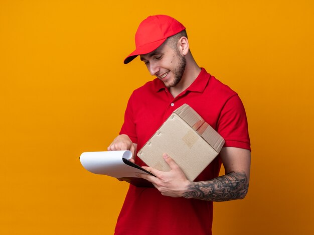 Foto gratuita repartidor joven complacido con uniforme con caja de sujeción de tapa mirando el portapapeles en la mano